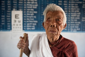 An elderly lady from Nepal who is blind stands in front of an eye chart. She is holding a walking stick.