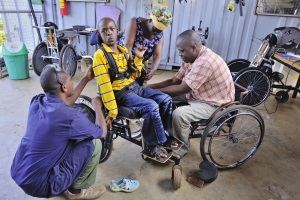 Picture of a teenage boy from Tanzania sitting in his wheelchair whilst it is being adjusted. There are two technicians carrying out the repairs, one of the technicians is also a wheelchair user. His mother is watching the process from behind.