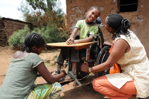 Picture of a girl from Tanzania who is sitting in a wheelchair fitted with a tray table outside a building.  She is smiling at the female CBR worker who is squatting beside her to her right. Her mother is squatting down in front of her adjusting a strap holding the child’s right foot in place on the wheelchair foot support. 