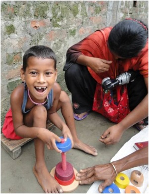 Picture of a girl from Bangladesh sitting on a stool, putting wooden shapes on a peg. The CBR worker is squatting beside her.