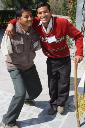 Picture of two smiling young men. One, who uses a walking stick , has his arm around his friend. His friend has no arms. 