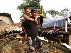 Picture of a woman from the Philippines supported on her father’s back. She has cerebral palsy. There is destroyed houses and debris in the background.  