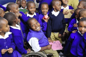 Picture of a young boy from Tanzania who uses a wheelchair. He is smiling and is surrounded by his classmates who are crowding around him.