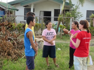 Picture of three students standing in front of a house that has been damaged by Typhoon Haiyan. They are talking to a visitor using sign language and an interpreter. There is a tree on the ground next to the students. 