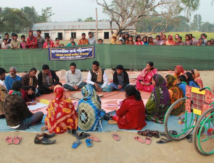 A Self-Help Group in Bangladesh is working together to prepare for and plan for disaster events that are inclusive and accessible. They are pictured sitting around a circle talking. 