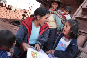 Picture of a girl from Peru sitting on a ladder outside of her house.  She is reading a book with the CBR worker. Her mother and baby sister are watching.