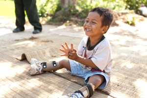 Picture of a smiling young boy from Cambodia sitting outside on a mat. He is wearing braces on both of his legs.