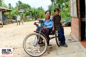 Sieng Sok Chann sits in her hand driven tricycle wheelchair outside her house with a large smile. Her young son leans against her shyly. There is a ramp leading to the doorway. In the background across the road are several more houses surrounded by trees, and a villager and small child walk are walking together along the road.