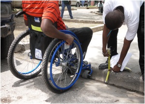 Picture of a man in a wheelchair stopped at a curb. Another man is bending over measuring the curb height with a tape measure.