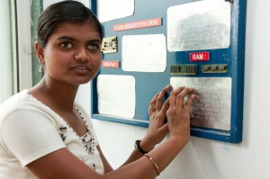 Picture of a young woman from India with a vision impairment reading Braille plates attached to the wall.  (The plates contain information about computer parts.)