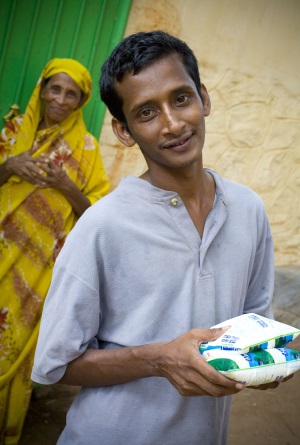 Picture of a man from India holding a packet of milk that is for sale. His mother is in the background looking on.